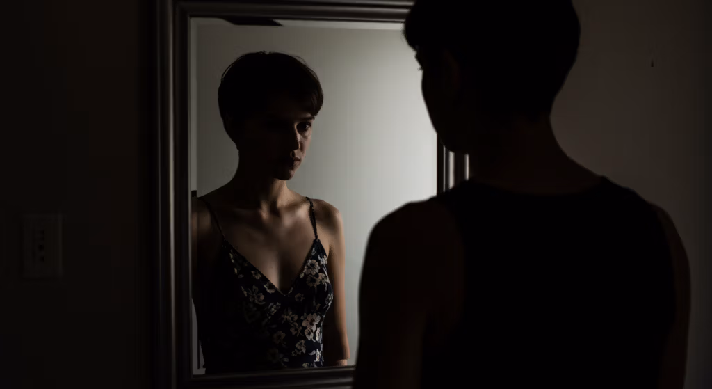 A person standing in front of a mirror, looking at their reflection. The person is wearing a sleeveless top with a floral pattern. The scene is dimly lit, creating a dramatic atmosphere with shadows and soft light.