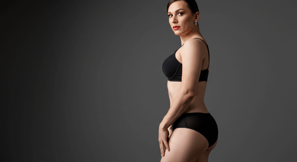 امرأة ترتدي ملابس داخلية سوداء تقف ويدها على وركها، وتقف بثقة أمام خلفية رمادية عادية.