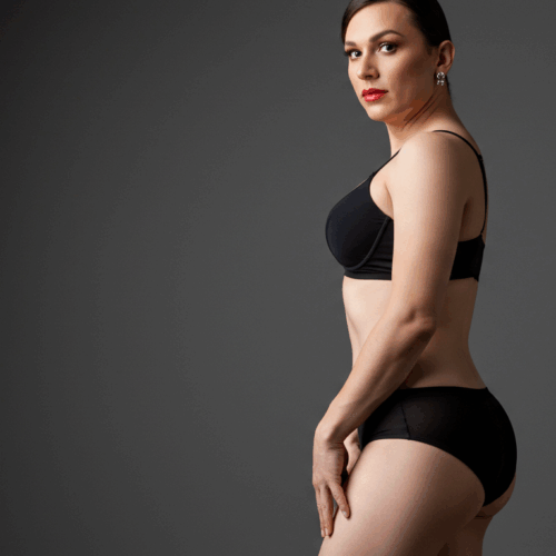 Une femme en sous-vêtements noirs debout avec une main sur la hanche, posant avec assurance sur un fond gris uni.