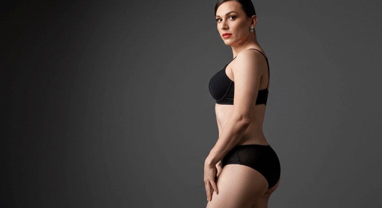 Siyah iç çamaşırlı bir kadın, bir elini kalçasına dayamış, düz gri bir arka plan önünde kendinden emin bir şekilde poz veriyor.