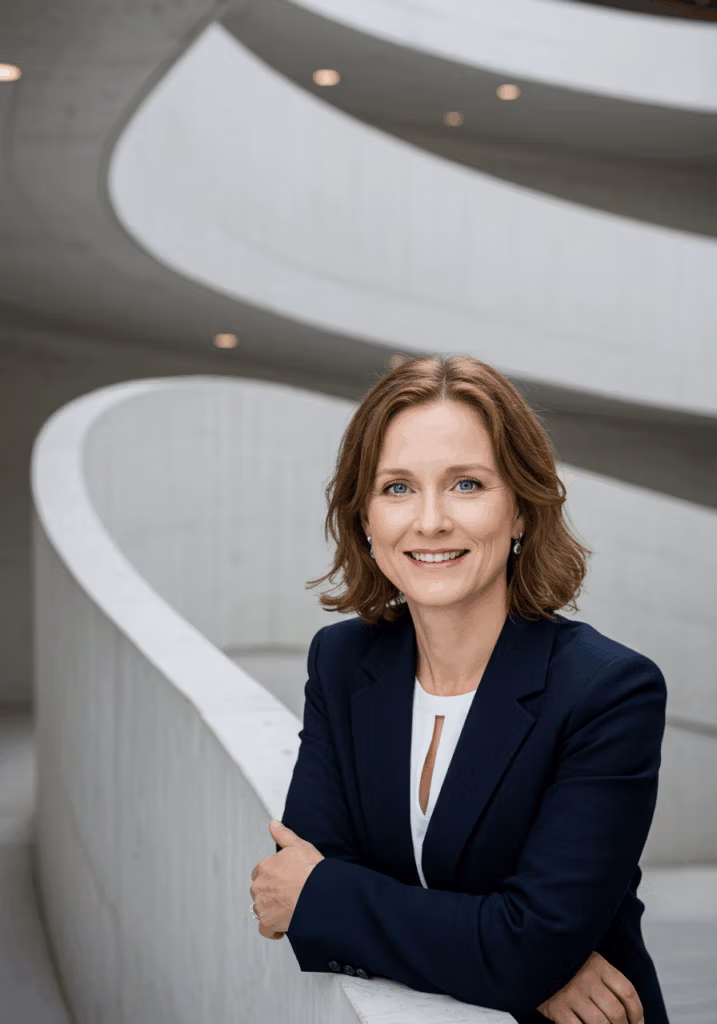 Retrato profesional de una mujer con cabello castaño corto, vestida con un blazer azul oscuro y una camisa blanca, apoyada contra una estructura curva de hormigón con un fondo arquitectónico moderno.