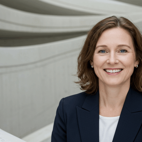 Une femme professionnelle aux cheveux bruns courts, portant un blazer foncé et une chemise blanche, debout devant un arrière-plan architectural moderne aux lignes courbes.