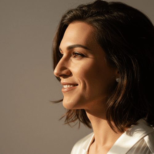 Een vrouw met kort, donker haar glimlacht en kijkt opzij. Ze draagt een lichtgekleurd topje en er valt zacht licht op haar gezicht.