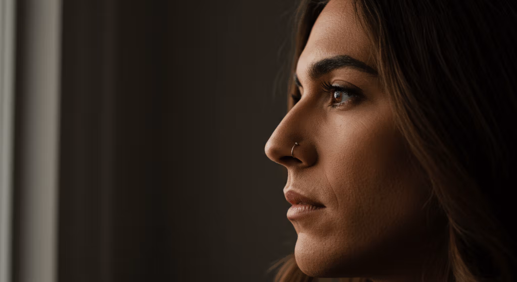 A close-up profile photo of a woman with a nose ring, looking to the side against a dark background.
