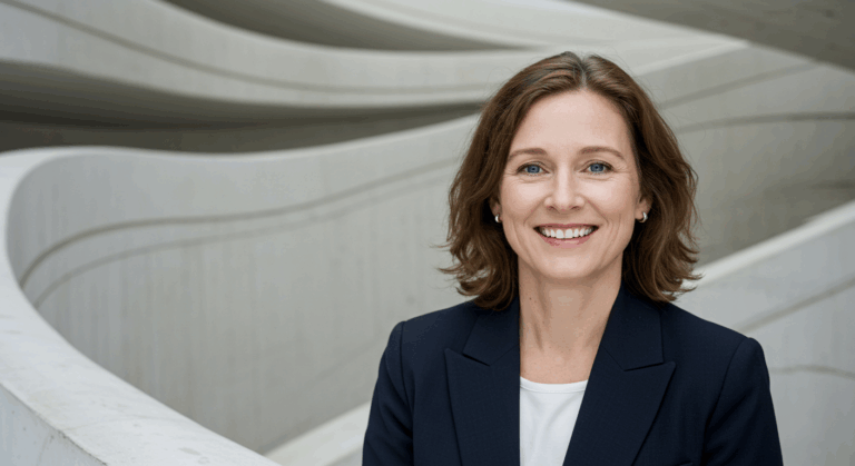 A professional woman with short brown hair, wearing a dark blazer and white shirt, standing in front of a modern architectural background with curved lines.