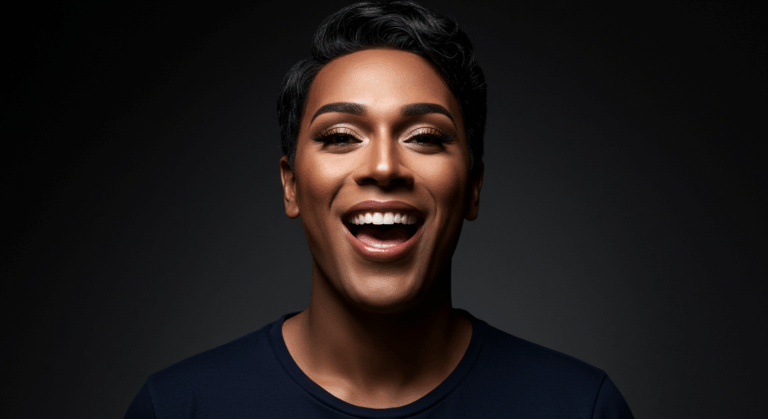 صورة مقربة لشخص مبتسم ذو شعر داكن قصير وبشرة فاتحة ورموش بارزة، يرتدي قميصًا أزرق غامقًا على خلفية سوداء.