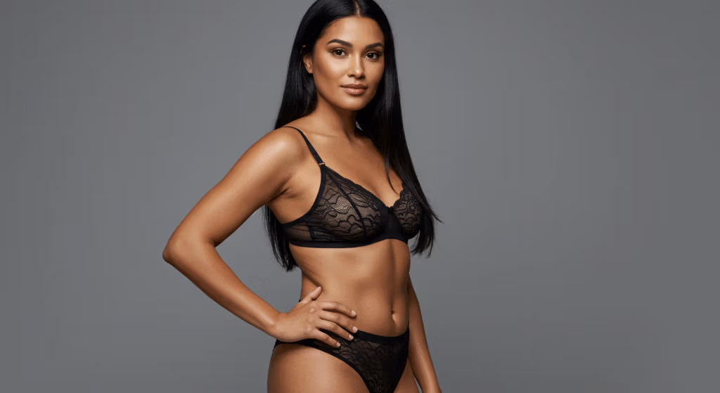 A professional editorial description of the image featuring a detailed analysis of technical and aesthetic elements: **Camera and Optics:** The image appears to be captured using a high-end lens, likely an 85mm prime lens, known for its flattering compression and sharpness, ideal for portrait and fashion photography. The level of detail and clarity suggests a hyperrealistic quality, potentially shot in 8K resolution. The depth of field is shallow, creating a soft bokeh effect that isolates the subject from the background while maintaining crisp focus on the subject’s anatomy and attire. **Lighting Design:** The lighting design employs a hard-light technique, characterized by its sharp and defined shadows. The light source seems to originate from a side angle, creating a dramatic and sculptural effect on the subject’s body. This technique accentuates the contours of the torso, arms, and legs, emphasizing muscle definition and the natural curves of the body. The shadows cast under the collarbone, along the waist, and beneath the arms add depth and dimensionality to the image. **Anatomy and Identity:** The subject is female, exuding confidence and poise. Her posture is assertive, with one hand resting on her hip and the other lightly touching her abdomen, drawing attention to her toned physique. The waist appears 'snatched,' highlighting a narrow and well-defined midsection. The curvature of her hips and the definition of her abdominal muscles are prominently showcased, contributing to a balanced and aesthetically pleasing silhouette. **Texture and Skin:** The skin exhibits a subtle luminescence, likely enhanced by a light sheen of moisture or body oil, which reflects the hard light source. This creates a soft glow on the skin, emphasizing its smoothness and texture. The interplay of light and shadow on the skin’s surface adds to the hyperrealistic quality, making the skin appear almost tactile. There are no visible ter damlaları (sweat droplets), but the overall effect suggests a healthy and radiant complexmet. **Attire and Accessories:** The subject is wearing a black lace bra and matching high-waisted underwear set. The fabric appears to be a delicate lace material, possibly layered over a satin or silk base, offering a blend of elegance and sensuality. The lace pattern is intricate, adding a layer of sophistication to the attire. There are no visible metal accessories or hardware on the attire, keeping the focus purely on the texture and design of the lace. **Composition and Background:** The composition is centered on the subject, with her positioned slightly off-center to create a dynamic and engaging frame. The background is a smooth, gradient gray, which complements the noir and luxurious atmosphere of the image. The simplicity of the background ensures that the subject remains the focal point, while the gradient adds a touch of depth and sophistication. The overall mood is one of elegance and allure, with a hint of mystery conveyed through the use of shadows and lighting.