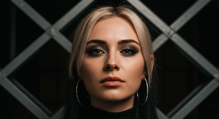 Sarı saçlı, koyu far ve kalın eyeliner içeren çarpıcı makyajlı, büyük halka küpeler takan, geometrik desenli koyu bir arka planın önünde duran bir kadının yakın çekim portresi.