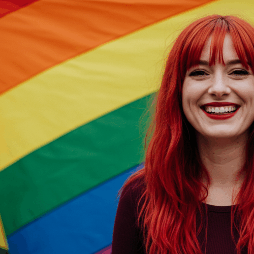 A smiling individual with vibrant red hair standing in front of a colorful rainbow flag, symbolizing LGBTQ+ pride and diversity.