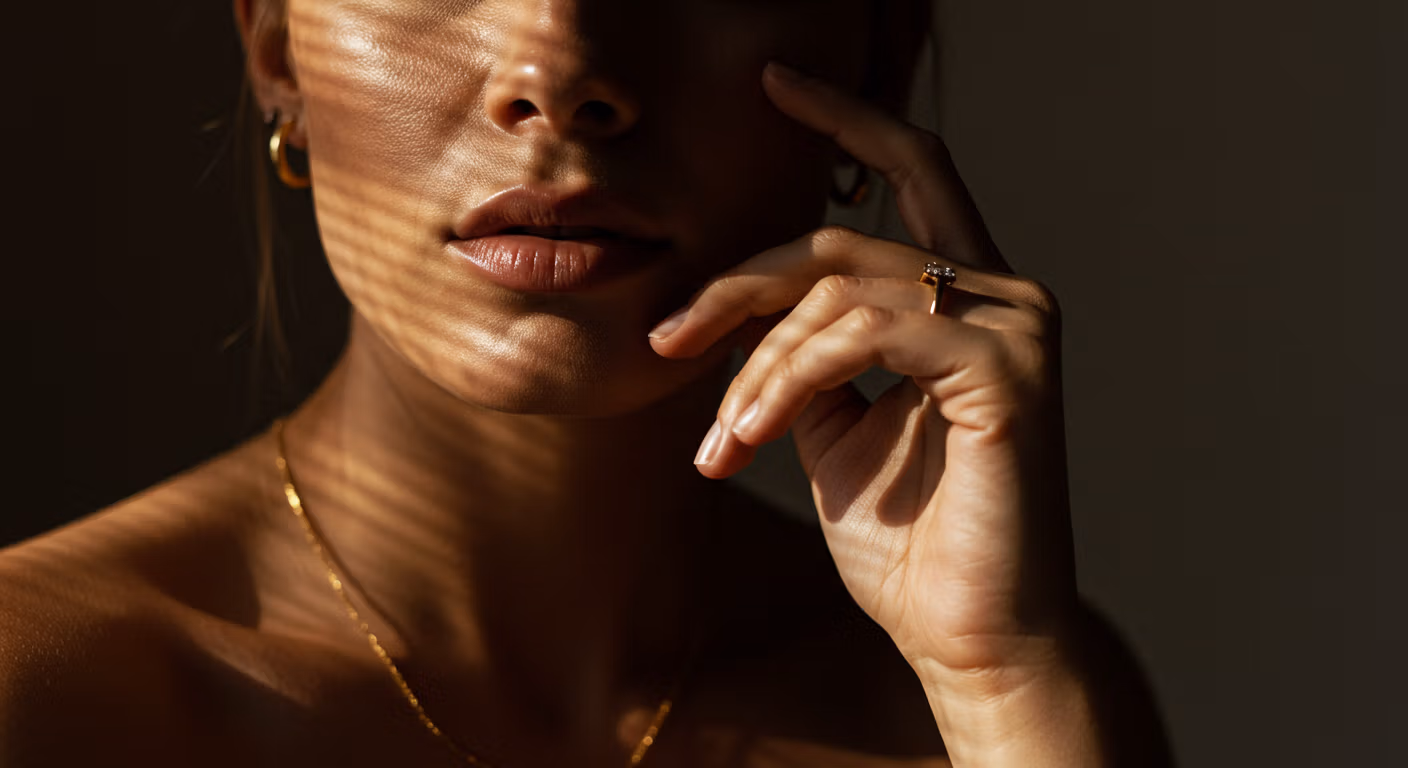 Gros plan du visage et de la main d'une femme portant des bijoux en or, notamment une bague et un collier. L'éclairage met en valeur ses traits et les reflets dorés des bijoux.