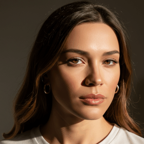 A woman with brown hair and gold earrings poses for a portrait in a room with natural light streaming in from a window.