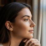 A young woman with dark hair is looking out of a window, with her hand resting on her chin, appearing thoughtful or contemplative.
