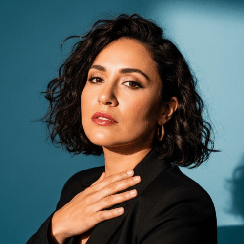 A woman with curly hair wearing a black top is posing for the camera with her hand on her chest. She has a serious expression on her face, and her shadow is visible on the wall behind her.
