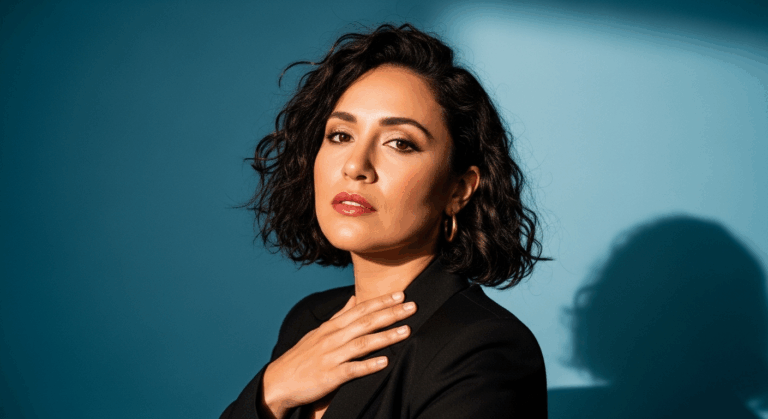 A woman with curly hair wearing a black top is posing for the camera with her hand on her chest. She has a serious expression on her face, and her shadow is visible on the wall behind her.