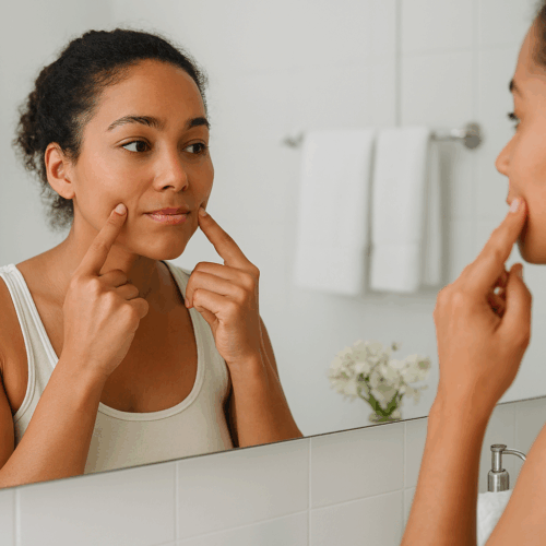 A woman is looking at her face in the mirror, examining her skin. She is touching her cheek with one hand while standing in a bathroom with white tiles and a vase of flowers on the counter.