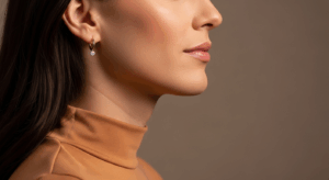 A close-up side profile of a woman wearing a high-necked, light orange top and small hoop earrings with a gemstone. She has smooth, dark hair and is posing against a plain brown background.