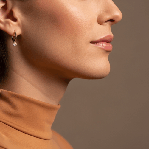 A close-up side profile of a woman wearing a high-necked, light orange top and small hoop earrings with a gemstone. She has smooth, dark hair and is posing against a plain brown background.