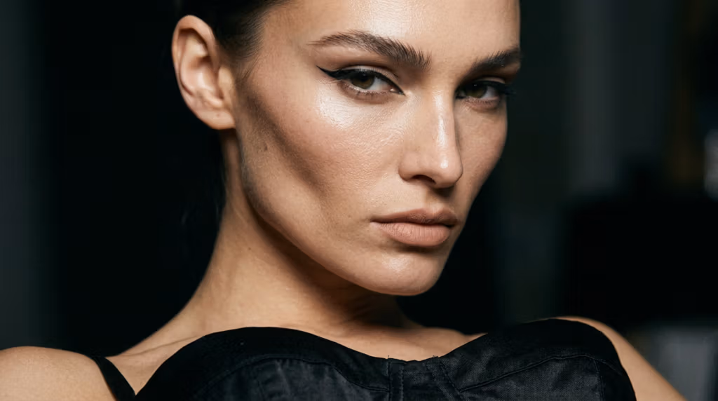 Close-up portrait of a woman with sharp winged eyeliner, defined contouring, and a natural makeup look, wearing a black top against a dark background.