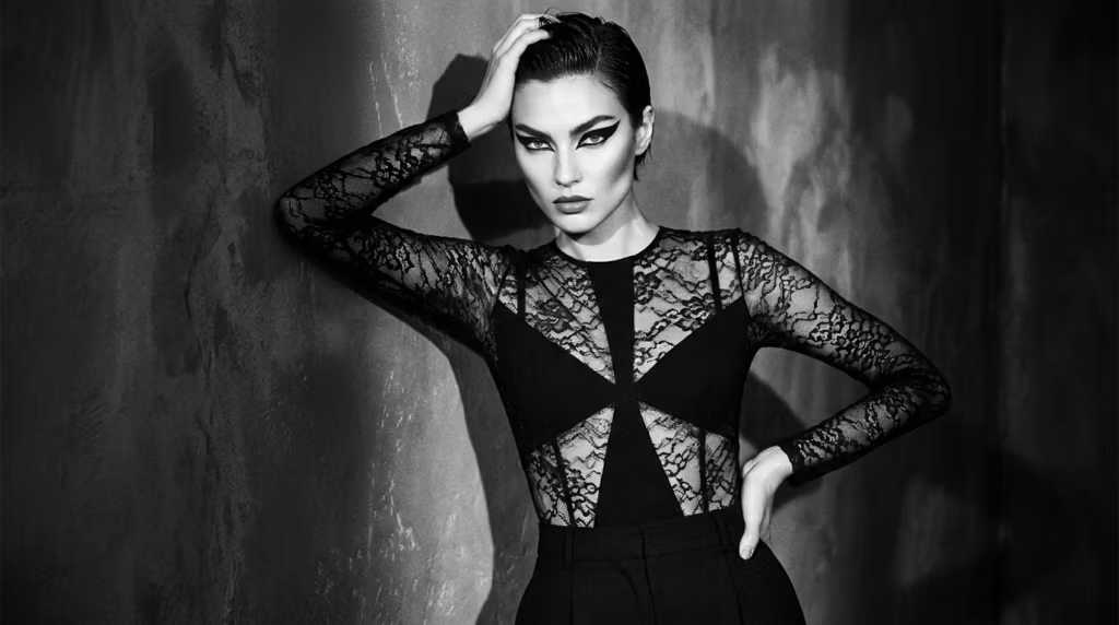 A high-fashion black and white portrait of a woman with bold graphic eyeliner, wearing a black lace-detailed top, posing confidently with a hand on her head against a dark, textured background.