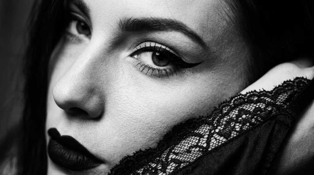 A black and white close-up portrait of a woman with dramatic eyeliner, dark lipstick, and intricate lace fabric near her face.