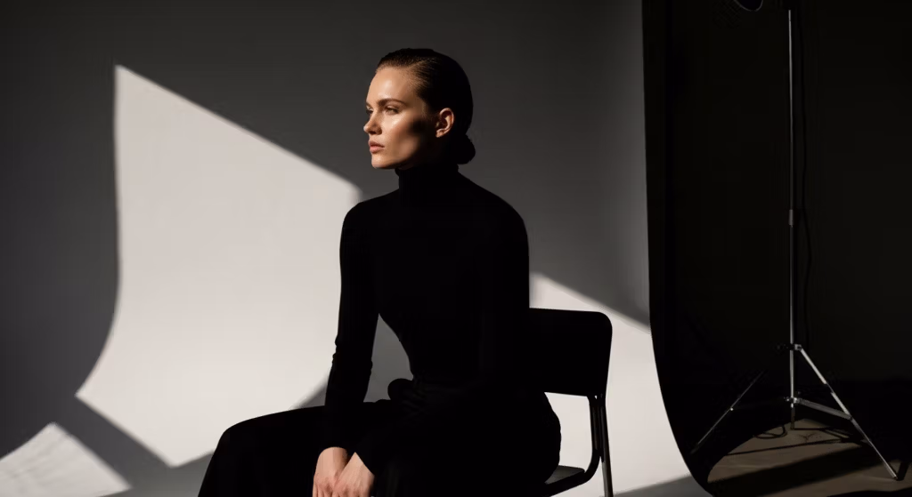 Portrait professionnel en studio d'une femme aux cheveux plaqués en arrière, portant un col roulé noir, assise sur une chaise devant un fond blanc, avec un éclairage latéral dramatique et un pied d'éclairage visible.