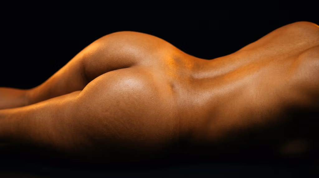 Gros plan sur le dos et les fesses lisses et toniques d'une personne à la peau éclatante, capturé en studio avec un éclairage latéral dramatique sur fond sombre.