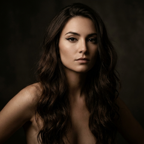 A professional studio portrait of a woman with long, wavy dark hair and elegant makeup, looking directly at the camera against a dark, moody background.