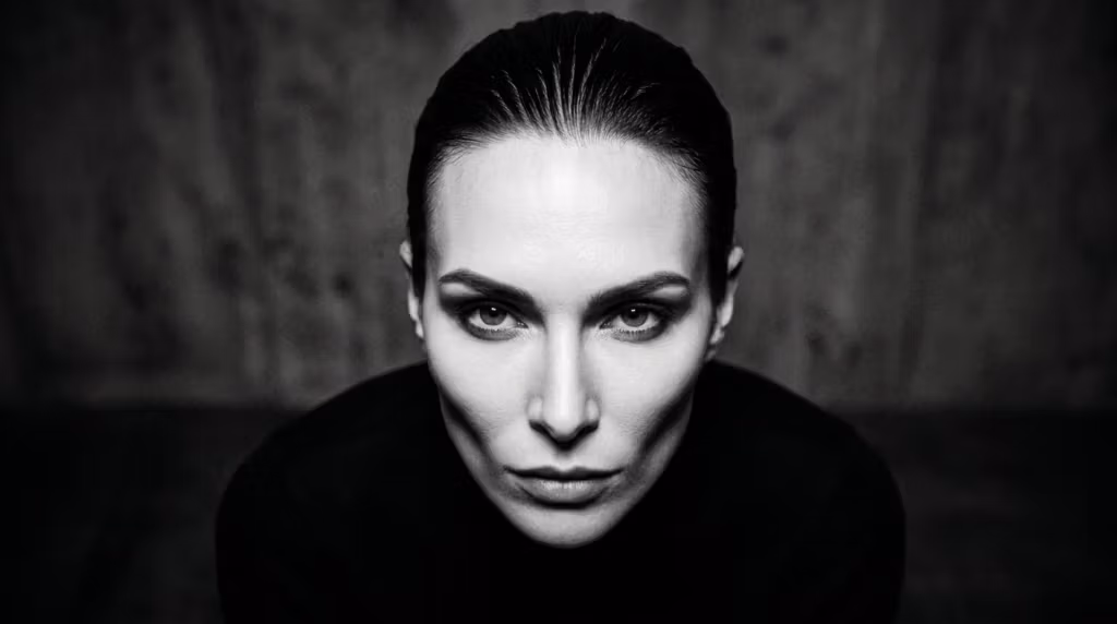 A striking, high-contrast, hyper-realistic 8K editorial portrait of a woman with a powerful, commanding gaze, captured with an 85mm prime lens. The lighting is masterfully executed with hard, directional side lighting that carves out her chiseled, high-fashion facial anatomy, emphasizing her sharp cheekbones and defined jawline with dramatic shadows. Her skin texture is rendered with impeccable micro-detail, displaying a subtle, natural luminescence and a sleek, dewy finish that catches the light. She is styled with her hair slicked back in a minimalist aesthetic, wearing a dark, matte-textured garment that keeps the focus entirely on her face. The background is a moody, textured, dark industrial surface, creating a sophisticated noir-inspired atmosphere that exudes strength, elegance, and raw intensity.