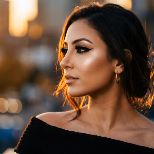 A hyper-realistic, 8K editorial portrait captured with an 85mm prime lens, focusing on a woman with refined facial features. The side-profile composition is illuminated by the dramatic, warm glow of a golden-hour sun, creating sharp contouring and a luminous highlight along her cheekbone and jawline. Her skin displays a flawless, radiant texture with subtle, dewy luminescence that catches the light. She is wearing a black, off-the-shoulder, textured garment that contrasts with the soft, out-of-focus urban background. Small gold hoop earrings add a touch of sophisticated hardware. The overall atmosphere is luxurious and cinematic, emphasizing an elegant, poised posture and meticulous makeup application.
