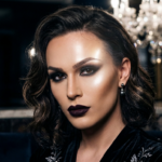 Captured with an 85mm portrait lens, this hyper-realistic 8k editorial headshot features a woman with a sharp, poised demeanor. The lighting is masterfully executed with a dramatic side-lit technique that carves out her refined facial structure, highlighting sculpted cheekbones and a defined jawline. Her skin displays a flawless, luminous texture, enhanced by expert contouring and subtle, radiant highlights that catch the light. She is adorned in a luxurious black velvet garment with intricate, shimmering metallic embroidery. The composition focuses on her piercing gaze and deep, matte plum-toned lips, set against a dark, moody background blurred by a soft-focus crystal chandelier, evoking a sophisticated, high-end noir ambiance.