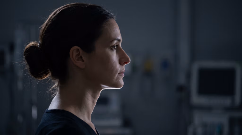 Ein hyperrealistisches, filmisches Porträt, aufgenommen mit einem 85-mm-Festbrennweitenobjektiv, das das elegante Profil einer Frau in den Mittelpunkt rückt. Die Komposition nutzt ein dramatisches Hell-Dunkel-Spiel: Eine scharfe, gerichtete Lichtquelle von hinten und oben erzeugt einen markanten Streiflichteffekt, der die Silhouette ihrer Kieferpartie und ihres Halses vor den tiefen, gedeckten Tönen der Umgebung hervorhebt. Die Bildqualität ist makellos und gibt jede Pore und jede subtile Hautstruktur in 8K-Auflösung gestochen scharf wieder. Ihr Ausdruck zeugt von stoischer Kontemplation, eingefangen in einer engen, redaktionellen Brustaufnahme. Der Hintergrund ist leicht unscharf und suggeriert durch die undeutlichen Umrisse medizinischer Monitore eine klinische oder sterile Umgebung, wodurch eine düstere, an Film Noir erinnernde Atmosphäre entsteht. Sie trägt ein schlichtes, dunkles, fein gestricktes Kleid, das in den Schatten verschwindet und so ihre anmutige Haltung und die Leuchtkraft ihrer Haut im weichen, stimmungsvollen Umgebungslicht vollends betont.