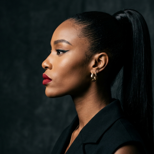 A hyper-realistic, 8k editorial portrait of a woman in profile, captured with an 85mm prime lens for razor-sharp clarity and flattering compression. The lighting is masterfully executed using a side-angle hard-light source, creating dramatic shadows that contour her high cheekbones, jawline, and the elegant slope of her neck. She features a statuesque, poised posture with refined, sculpted facial features. Her skin possesses a natural, subtle luminescence with a clean, smooth texture, highlighted by a bold red lip and sharp cat-eye eyeliner. She is dressed in a structured, high-quality black tailored garment, complemented by a minimalist gold hoop earring that catches the light. The composition is focused on the subject against a moody, textured charcoal-grey backdrop, evoking a sophisticated, modern noir aesthetic.