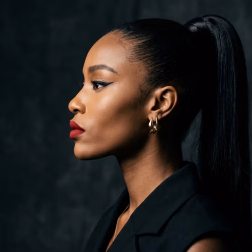 Ein hyperrealistisches 8K-Porträt einer Frau im Profil, aufgenommen mit einem 85-mm-Festbrennweitenobjektiv für gestochen scharfe Klarheit und schmeichelhafte Kompression. Die Beleuchtung ist meisterhaft mit einer seitlich einfallenden harten Lichtquelle umgesetzt, die dramatische Schatten erzeugt und ihre hohen Wangenknochen, ihre Kinnlinie und die elegante Halslinie betont. Sie hat eine statuenhafte, anmutige Haltung mit feinen, modellierten Gesichtszügen. Ihre Haut besitzt einen natürlichen, subtilen Schimmer mit einer reinen, glatten Textur, die durch einen kräftigen roten Lippenstift und einen präzisen Cat-Eye-Lidstrich hervorgehoben wird. Sie trägt ein strukturiertes, hochwertiges schwarzes Kleidungsstück, das durch einen minimalistischen goldenen Creolen-Ohrring, der das Licht einfängt, ergänzt wird. Die Komposition konzentriert sich auf das Motiv vor einem stimmungsvollen, strukturierten anthrazitgrauen Hintergrund und evoziert eine anspruchsvolle, moderne Noir-Ästhetik.