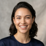 A high-resolution editorial portrait captured with an 85mm prime lens, showcasing a woman with a genuine, beaming smile. The lighting is soft and flattering, utilizing a multi-point studio setup that eliminates harsh shadows while emphasizing the natural contours of her face and subtle skin texture. Her skin appears fresh and natural, with a healthy, subtle glow and no artificial sheen or moisture. She is wearing a simple, dark navy blue satin top, complemented by delicate gold hoop earrings that add a refined touch. The composition is a classic head-and-shoulders shot, set against a neutral, textured grey fabric backdrop that lends a sophisticated, minimalist aesthetic to the image. The overall atmosphere is professional, approachable, and high-end, typical of modern commercial portrait photography.