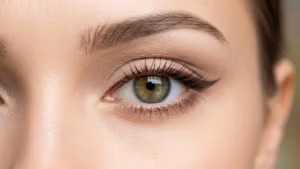 Eine sehr detaillierte redaktionelle Beschreibung des Bildes, das eine Nahaufnahme eines menschlichen Auges zeigt: **Kamera und Optik:** Das Bild wurde offenbar mit einem hochauflösenden Makroobjektiv aufgenommen, wahrscheinlich einem 85-mm- oder 100-mm-Objektiv, das für extreme Nahaufnahmen konzipiert ist. Der Detailgrad lässt auf eine hyperrealistische Qualität schließen, möglicherweise wurde das Bild in 8K-Auflösung aufgenommen, wodurch die feinen Texturen von Wimpern, Haut und Iris deutlich sichtbar werden. Die geringe Schärfentiefe erzeugt eine gestochen scharfe Fokussierung auf das Auge, während die Umgebung sanft verschwimmt. **Lichtgestaltung:** Die Beleuchtung im Bild ist weich und dennoch gerichtet und wurde wahrscheinlich durch eine Kombination aus diffusen und leicht angewinkelten Lichtquellen erzielt. Das Licht scheint von der Seite zu kommen und erzeugt subtile Schatten, die die Konturen der Augenlider und die Wölbung des Auges betonen. Das Fehlen harter Schatten deutet auf die Verwendung von Softboxen oder Diffusoren hin, um eine schmeichelhafte und gleichmäßige Lichtverteilung zu erreichen. Die Lichtreflexe im Auge deuten auf eine gut positionierte Hauptbeleuchtung hin und verleihen dem Bild Tiefe und Dimension. **Anatomie und Identität:** Das Auge der abgebildeten Person steht im Mittelpunkt des Bildes, ohne erkennbare Hinweise auf Geschlecht oder Körperhaltung. Die Nahaufnahme betont die natürliche Anatomie des Auges, einschließlich der Irisstruktur, der Glätte des Augenlids und der feinen Anordnung der Wimpern. Das Bild gibt keine Auskunft über weitere anatomische Merkmale wie Körperform oder Muskeldefinition. **Textur und Haut:** Die Haut um das Auge wirkt glatt und gut mit Feuchtigkeit versorgt, mit einem dezenten Schimmer, der das weiche Licht reflektiert. Schweiß oder Feuchtigkeitströpfchen sind nicht sichtbar, die Hautstruktur ist jedoch fein detailliert und zeigt natürliche Poren und feine Linien. Die dichten, definierten Wimpern verleihen dem Bild Realismus und Tiefe. Die Iris weist eine satte, natürliche Farbe mit feinen, scharf abgebildeten Mustern auf. **Kleidung und Accessoires:** Das Bild zeigt keine sichtbare Kleidung, Stoffstrukturen oder Accessoires. Der Fokus liegt allein auf dem Auge und seiner unmittelbaren Umgebung, wodurch jegliche Ablenkung durch äußere Elemente vermieden wird. **Komposition und Hintergrund:** Die Komposition ist eng gerahmt, wobei das Auge zentral positioniert ist, um die Aufmerksamkeit des Betrachters direkt auf seine Details zu lenken. Der Hintergrund ist bewusst unscharf, wodurch ein neutraler und unaufdringlicher Rahmen entsteht, der den Fokus auf das Auge verstärkt. Die Bildwirkung tendiert zu einer minimalistischen und klinisch-ästhetischen Ästhetik und betont Klarheit, Präzision und die natürliche Schönheit des menschlichen Auges. Das Fehlen von Kontext verleiht dem Bild eine zeitlose und universelle Qualität.