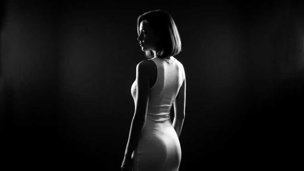 Descripción editorial profesional de la imagen: Un retrato en blanco y negro de alto contraste de una mujer con un vestido ajustado, que enfatiza la iluminación dramática, la definición anatómica y la textura lujosa. --- **Cámara y óptica:** La imagen parece haber sido capturada con un objetivo fijo de alta gama, probablemente un 85 mm f/1.4 o similar, conocido por su capacidad para producir imágenes nítidas y detalladas con poca profundidad de campo. Esta distancia focal es ideal para retratos, ya que ofrece un efecto de compresión favorecedor que acentúa los rasgos del sujeto. La calidad de la imagen sugiere una captura de alta resolución, posiblemente en 8K o con detalles hiperrealistas, lo que garantiza que cada textura y contorno se represente con precisión. La poca profundidad de campo aísla al sujeto del fondo, creando un enfoque visual impactante en su figura. --- **Diseño de iluminación:** La iluminación en esta imagen se caracteriza por una técnica de luz dura, que crea sombras y luces nítidas y bien definidas. La fuente de luz parece provenir de un ángulo lateral, probablemente de 45 grados o menos, lo que realza la tridimensionalidad de la figura. Esta iluminación lateral dramática acentúa los contornos de su cuerpo, en particular las curvas de su cintura, caderas y hombros, mientras proyecta sombras profundas que añaden una sensación de misterio e intensidad. El alto contraste entre luz y sombra enfatiza la cualidad escultórica de su anatomía, contribuyendo a una composición visualmente impactante. --- **Anatomía e identidad:** La retratada es una mujer con una postura segura y serena. Su postura es erguida, con una ligera curvatura en la espalda que acentúa la curvatura natural de su columna. La imagen resalta su anatomía femenina, en particular la definición de su cintura, descrita como "estrecha", que crea una silueta de reloj de arena. El juego de luces y sombras enfatiza aún más el volumen y la forma de sus contornos musculares y estructurales, incluyendo la definición de sus hombros, brazos y piernas. El efecto general es de elegancia y fuerza, combinando gracia con una presencia poderosa. --- **Textura y piel:** La piel en esta imagen exhibe una cualidad luminosa, con un brillo sutil que sugiere una ligera capa de humedad o aceite, lo que realza las propiedades reflectantes de la piel bajo la luz intensa. Si bien no hay gotas de sudor visibles, la textura de la piel parece suave pero detallada, con la luz captando los contornos naturales y las imperfecciones. La luminosidad de la piel añade una cualidad táctil a la imagen, haciendo que el sujeto parezca casi tangible. La captura de alta resolución asegura que los detalles finos de la superficie de la piel sean visibles, lo que contribuye al efecto hiperrealista. --- **Ropa y accesorios:** El sujeto lleva un vestido ajustado que parece estar hecho de una tela lujosa, probablemente satén o un material similar al satén. La tela se adhiere a su cuerpo, resaltando sus rasgos anatómicos y añadiendo sensualidad a la imagen en general. La textura del vestido refleja la luz sutilmente, contribuyendo a la cualidad luminosa de la piel. No hay accesorios metálicos ni herrajes visibles en el vestido, lo que permite que la atención se centre exclusivamente en la figura de la modelo y la textura de la tela. --- **Composición y fondo:** La composición de la imagen se centra en la modelo, con un fuerte énfasis en su silueta y el juego de luces y sombras. El fondo es oscuro y sin rasgos distintivos, creando una atmósfera sombría que contrasta marcadamente con la modelo iluminada. Este fondo minimalista garantiza que la atención del espectador se mantenga en la figura de la mujer y los intrincados detalles de la iluminación. El ambiente general de la imagen es de sofisticación e intensidad, evocando una sensación de lujo y elegancia atemporal. El uso del espacio negativo alrededor de la modelo realza aún más el efecto dramático, convirtiéndola en el punto focal indiscutible de la composición.