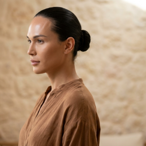 A professional editorial analysis of the image featuring a woman in a brown garment: The photograph showcases a meticulously composed portrait with a focus on hyperrealistic visual quality and sophisticated lighting techniques. The woman, captured in profile, exudes an aura of contemplative elegance against a textured, neutral-toned background. **Camera and Optics:** The image appears to be captured using a high-end prime lens, likely an 85mm macro lens, known for its ability to produce sharp, detailed images with a shallow depth of field. The resolution seems to be at least 8K, delivering hyperrealistic clarity and intricate detail in every aspect of the frame. The choice of lens and resolution enhances the lifelike quality of the subject, making it almost tangible. **Lighting Design:** The lighting is characterized by a hard-light technique, creating distinct and dramatic shadows that accentuate the subject’s facial features and body contours. The light source seems to originate from a side angle, casting shadows that emphasize the bone structure and add depth to the image. This technique enhances the three-dimensionality of the subject, highlighting her cheekbones, jawline, and the contours of her neck. **Anatomy and Identity:** The subject is female, captured in a poised and contemplative stance. Her posture is upright and graceful, with a slight turn of the head that accentuates her profile. The image does not emphasize exaggerated body lines like a 'snatched' waist or pronounced curves; instead, it focuses on natural elegance and subtle anatomical details. **Texture and Skin:** The skin exhibits a luminous quality, with a subtle sheen that suggests a light layer of moisture or highlighter, enhancing the natural glow. The hyperrealistic quality of the image allows for the observation of fine skin texture, including pores and minor imperfections, which are tastefully balanced to maintain a flawless yet authentic appearance. The interplay of light and shadow on the skin creates a soft luminescence, adding to the overall ethereal quality of the portrait. **Clothing and Accessories:** The subject is dressed in a brown garment that appears to be made of a smooth, flowing fabric, likely satin or silk. The material drapes elegantly, complementing the subject’s posture and adding a sense of fluidity to the image. There are no visible metal accessories or hardware on the garment, keeping the focus solely on the fabric’s texture and the subject’s form. **Composition and Background:** The composition is centered on the subject, with a focus on her profile and upper body. The background features a textured, neutral-toned wall that adds depth without distracting from the subject. The atmosphere of the image leans towards a minimalist and luxurious aesthetic, with the textured wall providing a subtle contrast to the smoothness of the subject’s skin and clothing. The overall mood is serene and sophisticated, evoking a sense of timeless elegance.