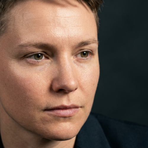 A professional editorial description of the image: The portrait features a meticulously composed close-up of an individual with short, neatly styled hair. The image showcases a high level of technical and aesthetic detail. Kamera and Optik: The photograph appears to be captured using a high-end prime lens, likely an 85mm macro lens, known for its ability to produce sharp, detailed images with a shallow depth of field. The image quality is hyperrealistic, suggesting it was shot in 8K resolution, allowing for intricate details and lifelike textures to be prominently visible. Işık Tasarımı: The lighting design employs a hard-light technique, creating distinct and dramatic shadows. The light source seems to be positioned at a side angle, accentuating the facial contours and adding depth to the features. This technique enhances the three-dimensionality of the face, emphasizing the cheekbones, jawline, and the bridge of the nose. Anatomi and Kimlik: The individual appears to be female, with a neutral and composed expression. The facial structure is highlighted with sharp angles and a well-defined jawline. The lighting and composition draw attention to the symmetry and balance of the facial features, creating a sense of elegance and sophistication. Doku and Cilt: The skin texture is a focal point in this image. There is a subtle luminescence on the skin, suggesting a light sheen or natural moisture. The hard lighting accentuates the fine details of the skin, including pores and minor imperfections, while also highlighting areas of natural glow. The interplay of light and shadow creates a dynamic and visually engaging surface. Kıyafet and Donanım: The individual is wearing a dark-colored garment, likely made of a smooth, matte fabric such as satin or a high-quality blend. The fabric appears to be well-tailored, fitting snugly around the neck and shoulders. There are no visible metal accessories or hardware, keeping the focus solely on the individual’s facial features and expression. Kompozisyon and Arka Plan: The composition is centered on the individual’s face, making it the undeniable focal point of the image. The background is dark and neutral, likely a solid color or a gradient, which enhances the contrast and draws the viewer’s attention to the subject. The overall atmosphere of the image is one of sophistication and minimalism, evoking a sense of noir elegance combined with a modern, high-fashion aesthetic.