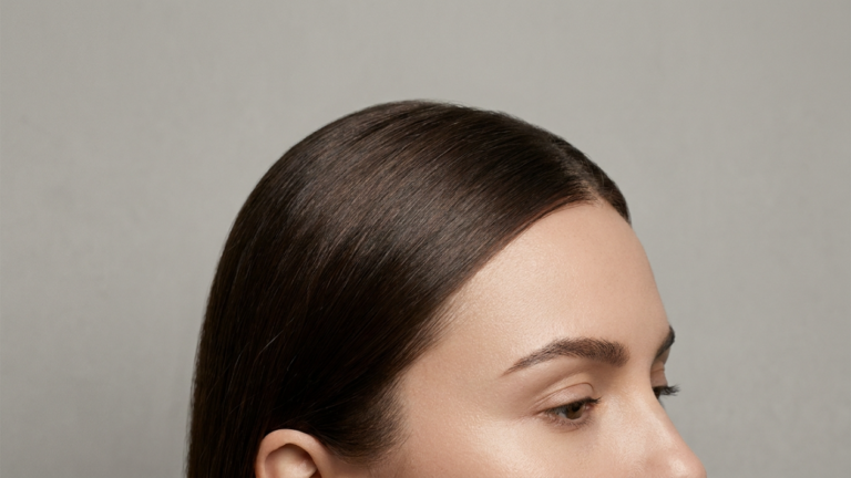 A hyper-realistic, 8k editorial portrait shot with an 85mm macro lens, capturing a close-up, profile-oriented view of a woman's upper face and sleek, dark hair. The lighting is soft and diffused, creating a clean, high-end studio aesthetic that highlights the natural, luminous texture of her skin and the precise grooming of her brows. The subject exudes a minimalist, sophisticated grace, with the focus razor-sharp on the skin's subtle pores and the individual strands of her polished, dark hair against a neutral, seamless grey background.
