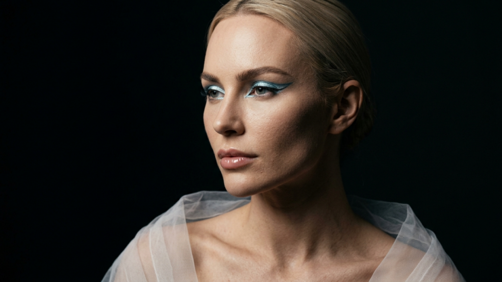 A high-fashion, hyper-realistic 8K editorial portrait of a woman with blonde hair pulled back, captured with a sharp 85mm prime lens to emphasize facial clarity. The lighting utilizes a dramatic side-angle hard light that sculpts her cheekbones and jawline, creating a sophisticated chiaroscuro effect against a deep, minimalist black background. Her complexion shows a subtle, healthy luminescence with refined skin texture. She wears a striking metallic blue graphic eyeliner that contrasts with the ethereal, translucent white tulle draping around her shoulders. Her expression is poised and serene, highlighting a sculptural beauty aesthetic that blends avant-garde makeup with a clean, luxury editorial style.