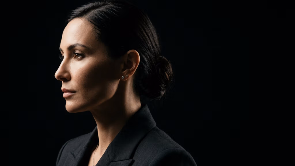 Un retrato editorial profesional y cinematográfico de una mujer, capturada en un perfil nítido, que demuestra la alta calidad de la fotografía DSLR con la claridad de un objetivo fijo de 85 mm. La iluminación está magistralmente ejecutada mediante una técnica de luz lateral direccional y dramática, creando un marcado contraste que esculpe sus rasgos faciales, resaltando el puente de la nariz, el pómulo y la mandíbula sobre profundas sombras negras. Su piel posee una textura natural y refinada con una luminosidad sutil y saludable, libre de imperfecciones. Irradia un aire de sofisticada autoridad, vestida con un blazer estructurado en tono carbón con solapas impecables. Su cabello oscuro está recogido en un moño pulido y elegante, que enfatiza una silueta definida. La composición es minimalista y centrada, utilizando el espacio negativo del fondo negro profundo para evocar una atmósfera de lujo y una intensidad profesional moderna de inspiración noir.