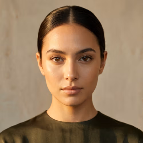 Een hoogwaardig redactioneel portret van een vrouw met een serene, symmetrische gelaatsstructuur, vastgelegd met een professionele 85mm-lens die een geringe scherptediepte creëert en scherpe details van 4K-kwaliteit benadrukt. De belichting is zacht maar gericht, doet denken aan het gouden uur, en werpt subtiele schaduwen die haar elegante kaaklijn en verfijnde gelaatstrekken accentueren. Haar huid heeft een natuurlijke, gehydrateerde gloed met een subtiele glans die het licht weerkaatst op de brug van haar neus en jukbeenderen. Haar haar is strak naar achteren gekamd in een minimalistische stijl, wat haar evenwichtige houding benadrukt. Ze draagt een kledingstuk van zwaar, olijfgroen satijn of zijde, dat een subtiele, verfijnde glans heeft. De achtergrond is een gedempte, getextureerde neutrale muur, die een minimalistische, eigentijdse sfeer creëert en de volledige aandacht vestigt op haar kalme, directe blik.
