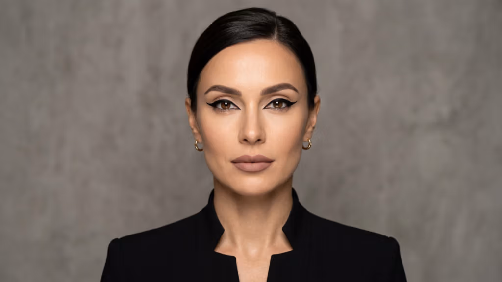 Ein professionelles, hochwertiges Studio-Porträt einer Frau, aufgenommen mit der gestochen scharfen Klarheit eines 85-mm-Porträtobjektivs, charakteristisch für Premium-DSLR-Fotografie. Die Beleuchtung ist meisterhaft: Weiches, diffuses Studiolicht erzeugt einen ausgewogenen, schmeichelhaften Glanz auf ihren symmetrischen Gesichtszügen und betont ihre feine Knochenstruktur und makellose Hautstruktur mit einer natürlichen, subtilen Leuchtkraft. Die Frau posiert mit einem selbstbewussten, direkten Blick und strahlt Eleganz und Selbstbewusstsein aus. Ihr Teint ist makellos, matt und wirkt gleichzeitig natürlich frisch. Sie trägt einen strukturierten, minimalistischen schwarzen Blazer mit einem klaren, eleganten Ausschnitt, der ihre Silhouette perfekt in Szene setzt. Zarte goldene Creolen verleihen dem Look einen Hauch von Luxus. Die Komposition ist ein zentriertes, enges Porträt vor einem strukturierten, neutralen Hintergrund in Betonoptik. So entsteht eine moderne, redaktionelle Atmosphäre, die von unaufdringlicher Eleganz und professioneller Souveränität geprägt ist.