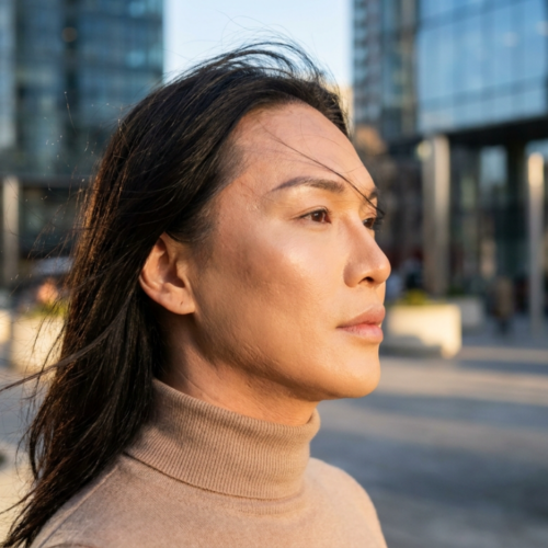 Un portrait éditorial haute résolution, réalisé avec un objectif fixe de 85 mm, révèle une personne transgenre dans une pose sereine et contemplative. L'image bénéficie d'une netteté professionnelle digne d'un reflex numérique, avec une faible profondeur de champ qui floute délicatement l'arrière-plan architectural urbain et moderne. La lumière naturelle latérale, typique de l'heure dorée, offre un éclairage subtil, mettant en valeur les pommettes hautes et les traits fins du visage, grâce à des ombres naturelles et subtiles. La peau, lisse, éclatante et hydratée, capte la lumière et lui confère une luminosité discrète. La personne porte un pull à col roulé beige en maille fine, qui s'harmonise avec la palette de couleurs chaudes. L'ensemble de la composition évoque une atmosphère sophistiquée, moderne et sereine, capturant un moment d'introspection paisible au cœur de la ville.