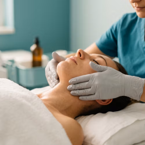 Een hogeresolutiefoto in redactionele stijl, gemaakt met een 85mm macrolens, die professionele huidverzorging en klinische luxe benadrukt. De compositie toont een cliënt die op haar rug ligt en een therapeutische gezichtsmassage krijgt van een therapeut. De belichting is zacht en diffuus, typisch voor een luxe medische spa, en werpt subtiele, natuurlijke schaduwen die de serene gelaatstrekken en ontspannen nek van de cliënt accentueren. De cliënt heeft een natuurlijke, gehydrateerde huidtextuur met een subtiele glans, wat duidt op vitaliteit na de behandeling. De therapeut draagt een professioneel turquoise scrubshirt en steriele lichtgrijze nitrilhandschoenen die een scherp, schoon contrast vormen. De achtergrond is licht onscherp en toont een steriele, rustige klinische omgeving met netjes opgestapelde witte handdoeken, wat een gevoel van kalmte, reinheid en luxe wellness oproept. De esthetiek is schoon, professioneel en verfijnd, en benadrukt de tactiele ervaring van de massage.