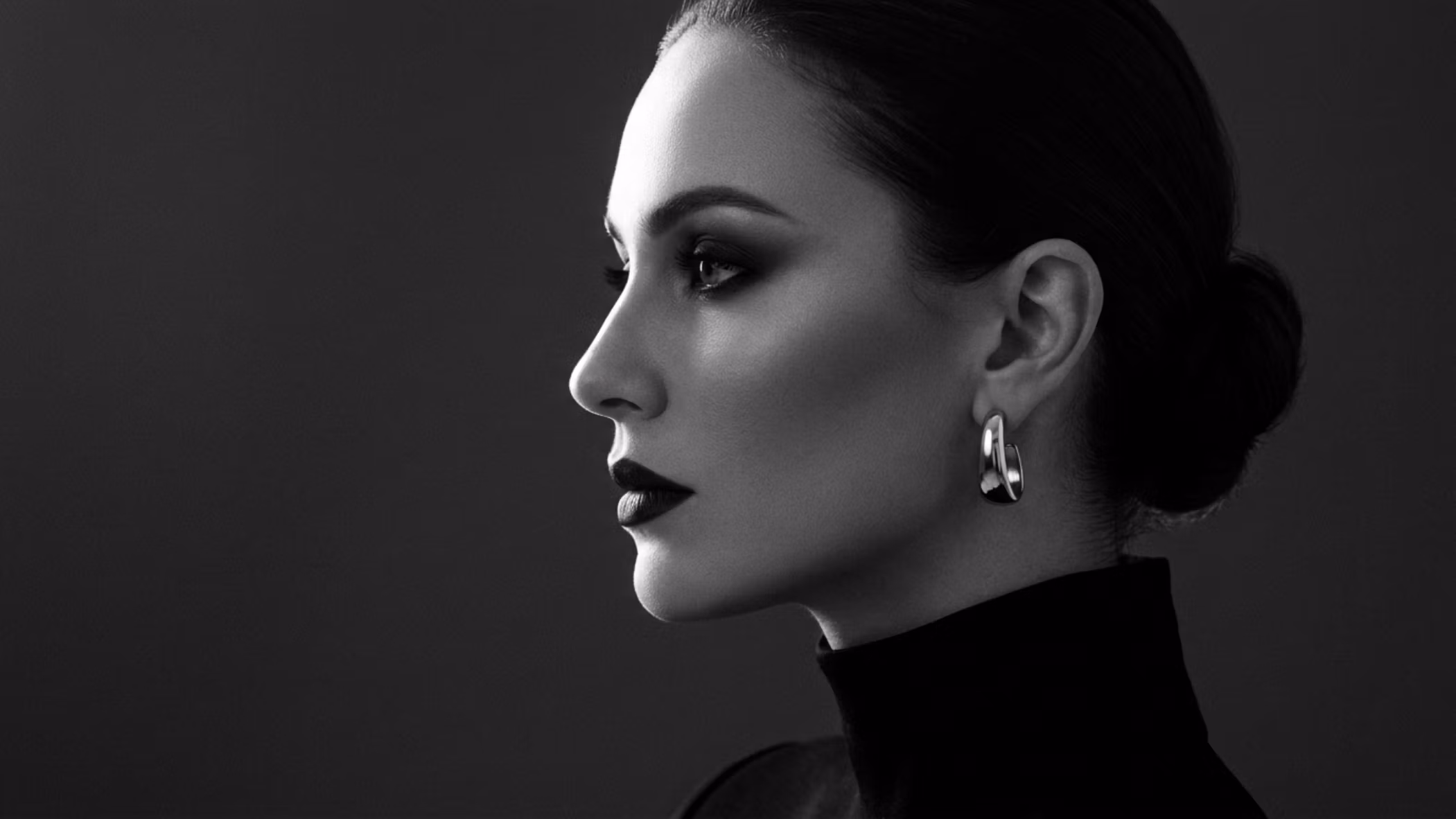 Un impactante retrato de estudio en blanco y negro de alto contraste, capturado de perfil con nitidez. Tomada con un objetivo fijo de 85 mm para una compresión óptima del retrato y la claridad de alta resolución de una cámara réflex digital, la imagen exhibe una sofisticada iluminación de estilo noir. La dramática iluminación de contorno lateral resalta la elegante estructura ósea de la mujer, acentuando su mandíbula definida, su nariz estilizada y su estética minimalista y refinada. Su tez luce un acabado mate y suave que interactúa maravillosamente con la luz, transmitiendo una sensación de serena luminosidad. Viste una prenda negra de cuello alto, con una sutil textura, combinada con un singular pendiente de aro de plata pulida que capta la luz como un refinado punto focal. La composición es limpia y minimalista, sobre un fondo oscuro y uniforme que enfatiza su expresión serena y contemplativa, así como la calidad atemporal y de alta costura de la pieza.