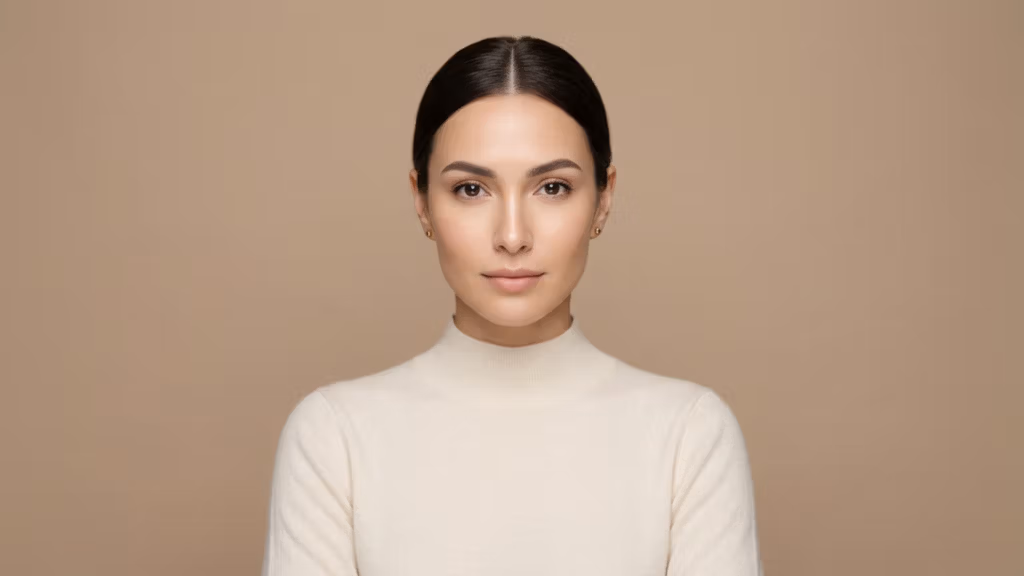 Retrato editorial de alta resolución, capturado con un objetivo fijo de 85 mm, que muestra a una mujer con una estructura facial refinada y simétrica, y una postura serena y centrada. La iluminación es suave y difusa, característica de la fotografía de estudio profesional, creando delicados contornos en sus pómulos y mandíbula sin sombras marcadas. Su piel luce un brillo natural e hidratado con una sutil luminosidad, que refleja una estética limpia y refinada. Viste un top de punto color crema de cuello alto con una textura delicada y suave que complementa el cálido fondo beige monocromático. La composición general es minimalista y sofisticada, enfatizando su mirada serena y sus rasgos equilibrados, plasmados con la nitidez y profundidad de color típicas de las imágenes DSLR de alta gama.