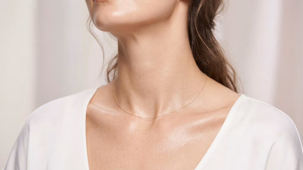 This high-definition, editorial-style close-up portrait, captured with an 85mm macro lens, epitomizes refined DSLR photography with its crisp 4K resolution and shallow depth of field. The lighting is masterfully soft and diffused, emanating from the side to create gentle, flattering shadows that accentuate the graceful contours of the subject’s neck and collarbone. The subject, a woman with an elegant, upright posture, exudes a serene and sophisticated air. Her skin possesses a natural, healthy luminosity, subtly enhanced by a faint, dewy sheen that catches the light with micro-reflective clarity. She is adorned in a minimalist, soft-textured white garment, complemented by a delicate, thin gold chain necklace that rests elegantly against her skin. The composition is tight and focused, set against a clean, neutral, and out-of-focus background that contributes to a minimalist, luxury aesthetic, placing full emphasis on the organic beauty and subtle textures of the subject.