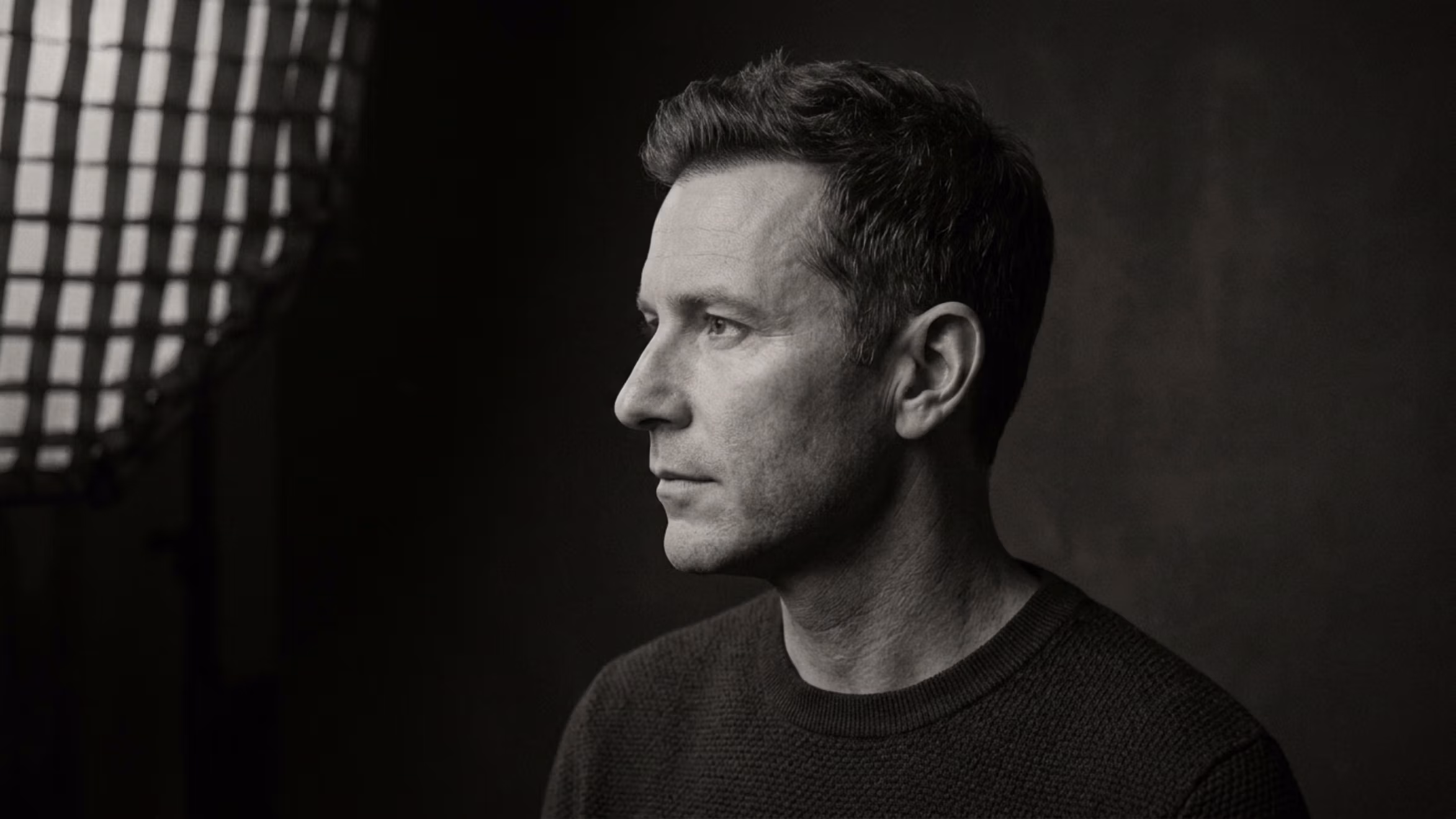 A sophisticated black-and-white editorial portrait of a middle-aged man, captured with the crisp precision of an 85mm prime lens on a high-resolution DSLR, showcasing impeccable detail. The lighting is masterfully executed using a side-lit softbox with a grid, creating dramatic chiaroscuro that carves out the subject's angular facial features—prominent brow, sharp jawline, and defined nose—with cinematic depth. The subject, a man with a composed, stoic expression, is positioned in a classic profile, conveying a sense of quiet authority. His skin texture is rendered with raw authenticity, revealing natural pores and subtle skin tone variations, highlighted by the soft luminescence of the directional light. He is dressed in a textured, dark-knit sweater that provides a tactile contrast to the smooth, muted backdrop. The overall composition is intimate and refined, reminiscent of classic studio portraiture, with the visible edge of the studio lighting gear in the upper left corner adding a touch of behind-the-scenes professional grit to the otherwise elegant noir-inspired aesthetic.