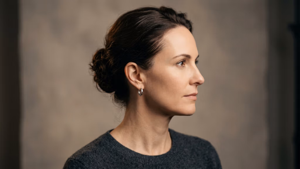 A professional, high-resolution DSLR studio portrait captured with an 85mm prime lens, emphasizing a refined aesthetic. The lighting is soft yet directional, striking from the side to subtly contour the subject's jawline and facial structure. The subject, a woman with a poised, profile-focused posture, wears her dark hair in a clean, sophisticated bun. Her skin texture is natural and pristine, reflecting a soft, healthy luminescence without artificial moisture. She is dressed in a minimalist, charcoal-gray knit sweater, accessorized only with a small, polished silver hoop earring. The composition is a tightly framed headshot, set against a neutral, mottled gray studio backdrop that evokes a timeless, editorial atmosphere. The overall image quality is exceptional, showcasing sharp focus on the eye and skin details, characteristic of high-end commercial portraiture.